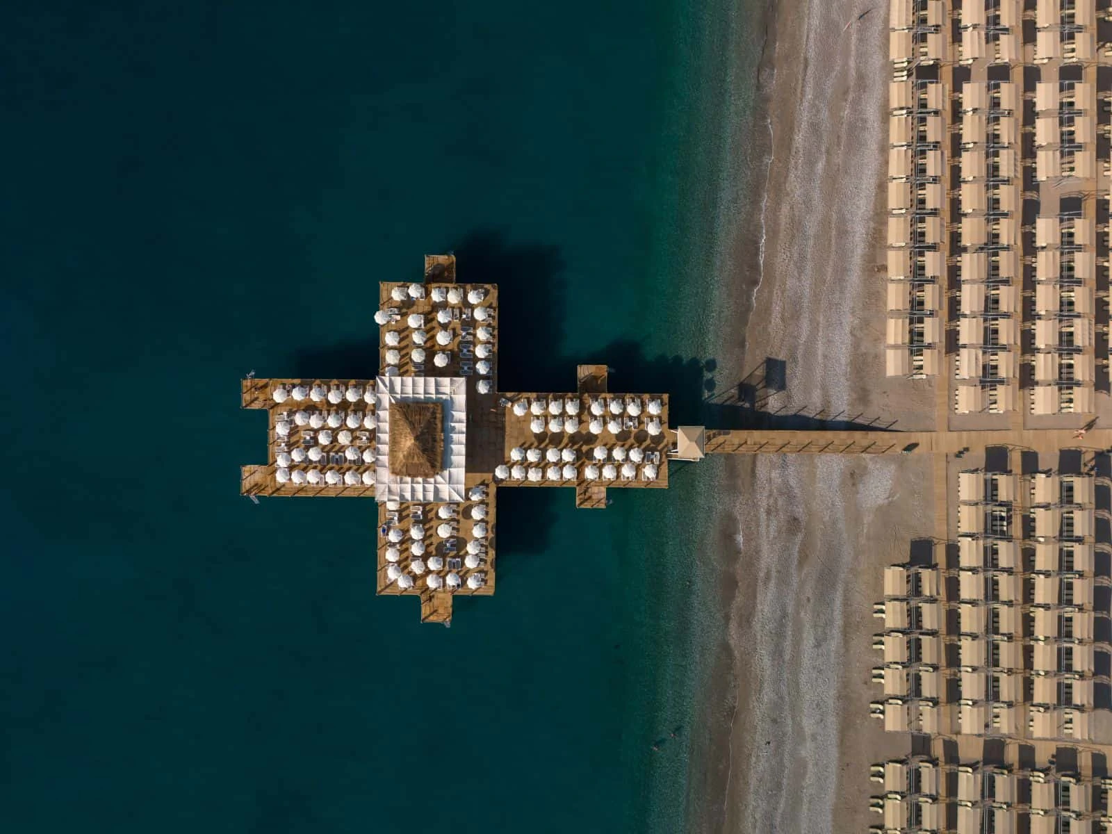 Luftaufnahme eines Badestegs mit Sonnenschirmen im türkisen Meer, daneben der Strand